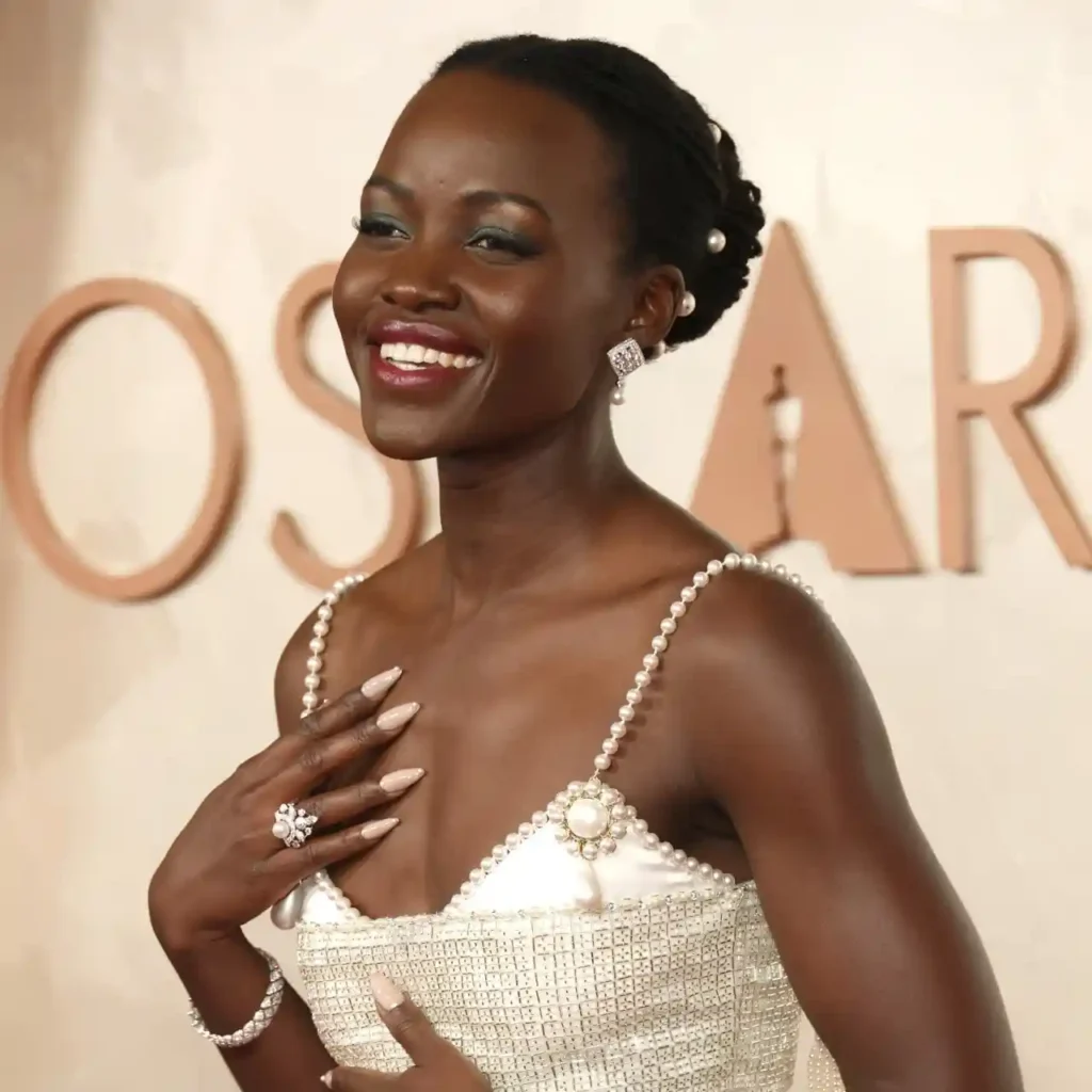 A smiling woman in a stylish, pearl-embellished dress poses gracefully at an awards event, showcasing exquisite jewelry and a sophisticated look.