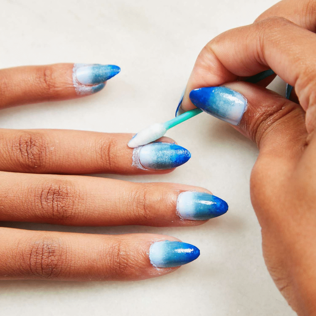 How to Master the Perfect Ombre Nail at Home 3 A close-up of a hand with beautifully painted blue ombre nails, as someone uses a cotton swab to clean up around the nail edges.