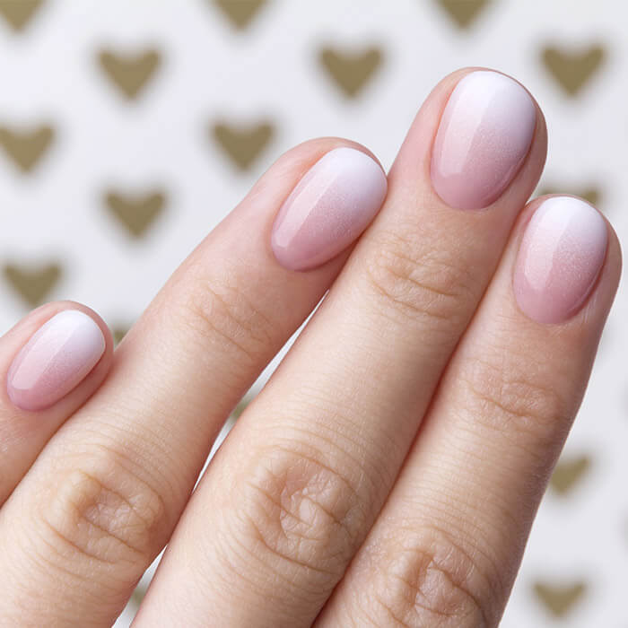 How to Master the Perfect Ombre Nail at Home 5 Close-up of a hand with elegant ombre nails featuring a gradient from soft pink to white, set against a background of gold hearts.